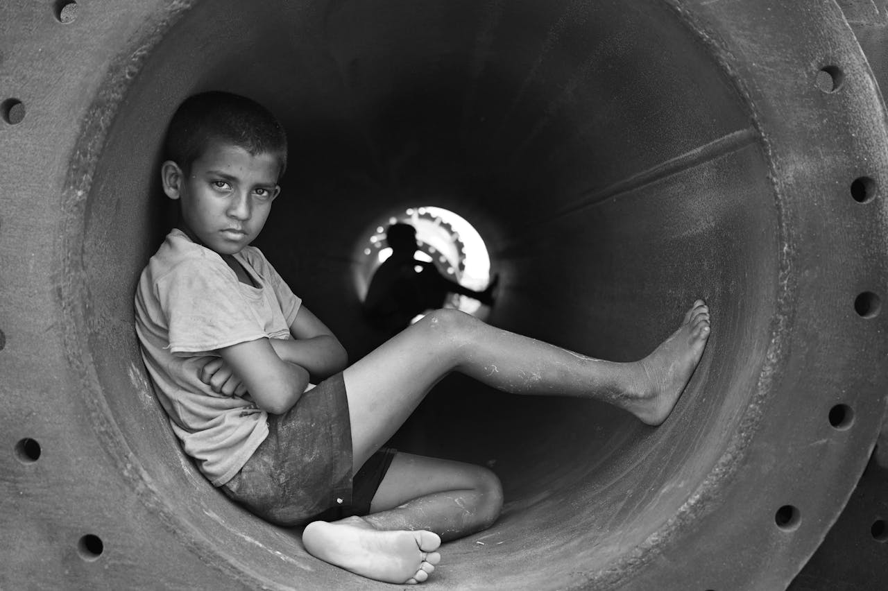 A contemplative young boy sits barefoot in a large industrial pipe in black and white.