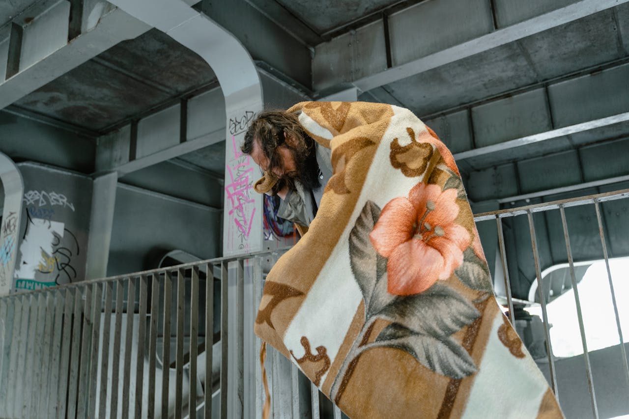 A bearded man wrapped in a floral blanket stands beneath an urban overpass.